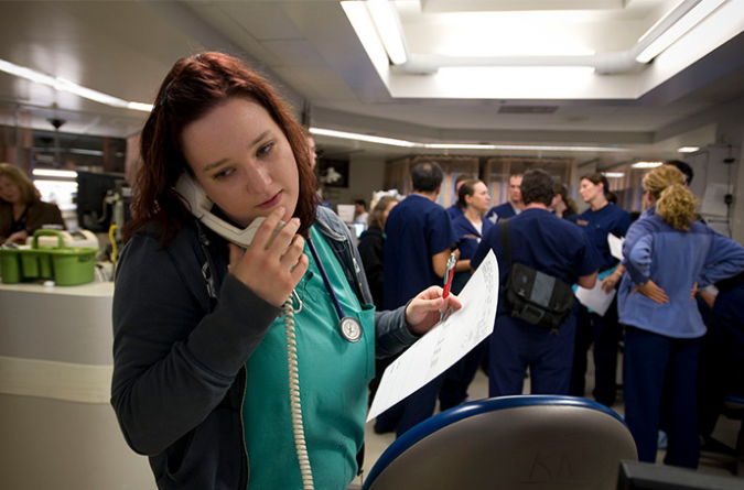 A nurse in an emergency department holds a phone in one hand and a clipboard in the other hand.