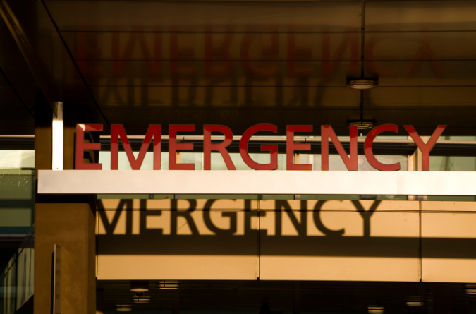 An "Emergency" sign is seen above a door as the letters are reflected in a window.