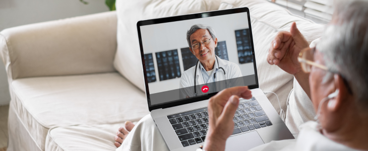 An older man sits on a couch with a laptop on his lap while on a video call with a doctor on the screen.