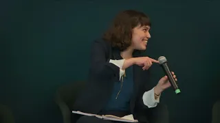 A woman in a dark coat smiles and looks to the side with a microphone in her hand.