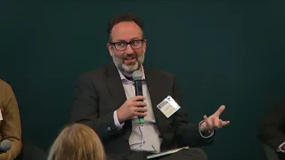 A man with a beard and a dark suit speaks into a microphone.