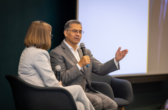 A seated man in a suit speaks into a microphone next to a woman in a suit.