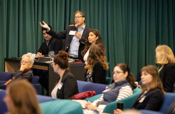 A man in a suit stand in an audience while speaking into a microphone. 