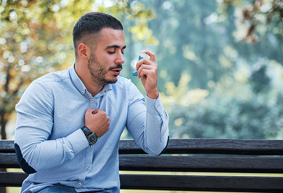 Man sitting on a park bench holding his chest as he’s about to use an inhaler.