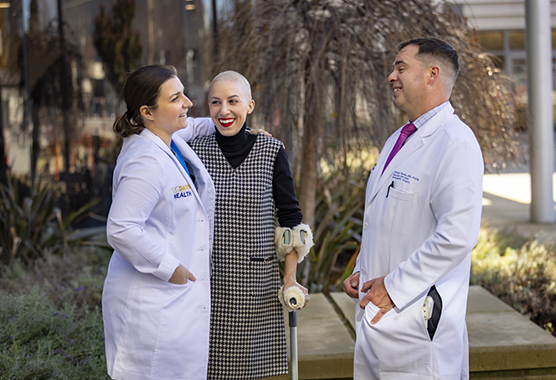 Female patient with shaved head smiling standing with a crutch alongside female and male health care providers outdoors.