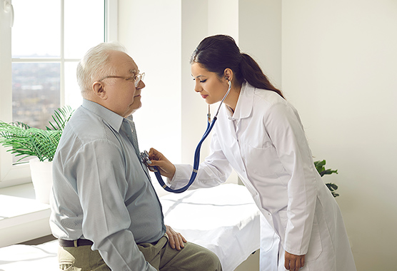 Older man sitting while female health care provider listens to his heart with stethoscope.