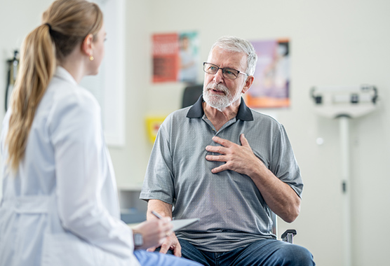 Older man sitting with his hand on his chest talking to a female health care provider in a clinic.