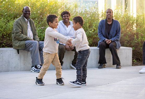 Two young boys holding hands and playing with three smiling adults watch