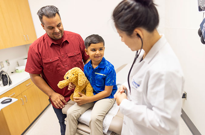 Young boy holding a teddy bear while sitting on exam table with his father and a female health care provider next to him