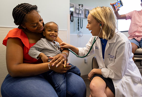 Young boy sitting on his mom’s lap smiling as female health care provider listens to his heart with a stethoscope