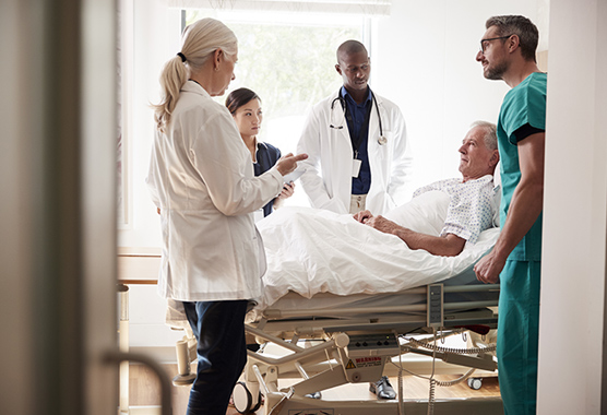 Older man in a hospital bed with health care professionals gathered around talking to him.