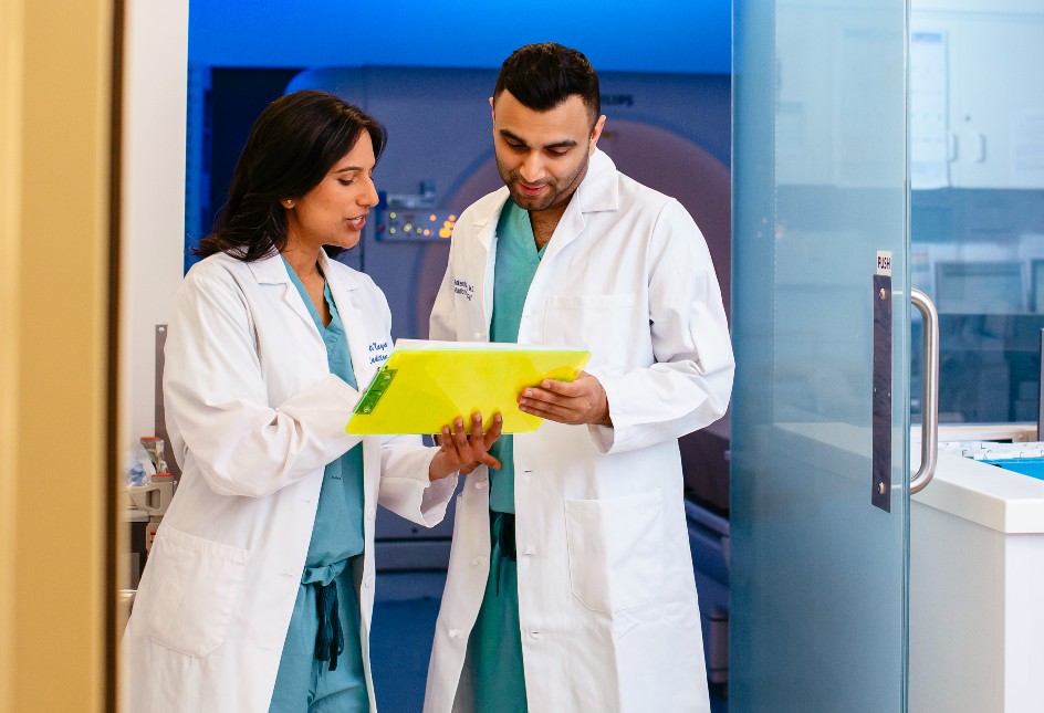 Two doctors reviewing documents on a yellow clipboard in front of a diagnostic imaging machine