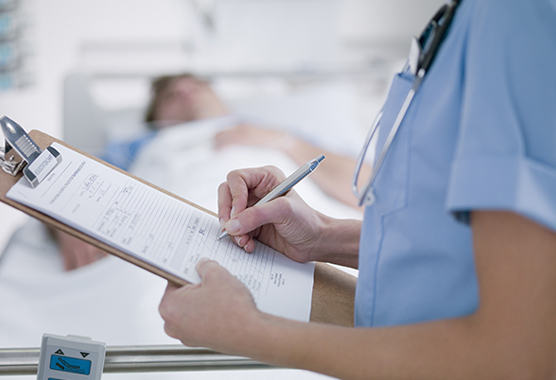 Health care provider writing on a clipboard with a patient laying on a hospital bed in the background.