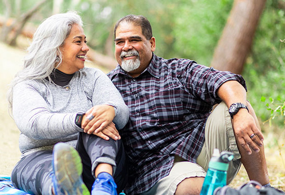 Older man and woman smiling sitting on a picnic blanket outside.