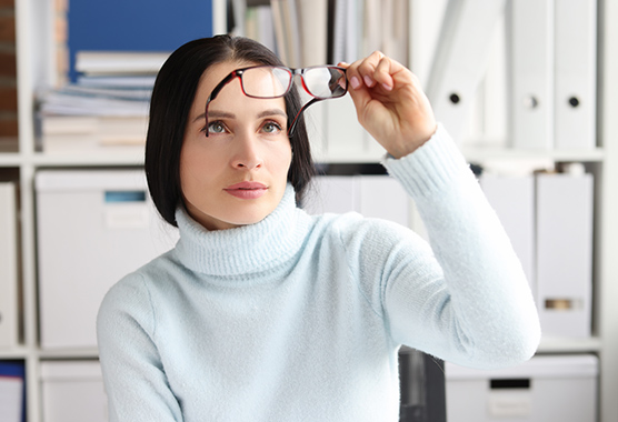 A woman holds her glasses away from her face and looks through the lens.