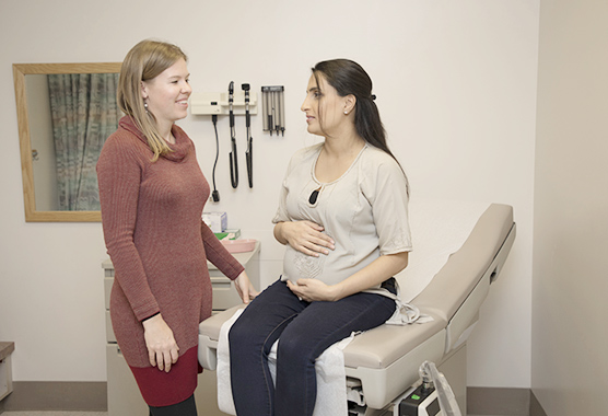 Pregnant woman sitting on an exam table holding her belly while talking to health care professional.