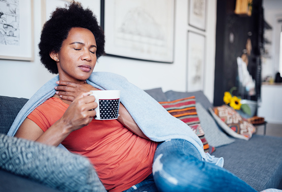 Woman sitting on a couch holding a coffee mug and touching her throat.