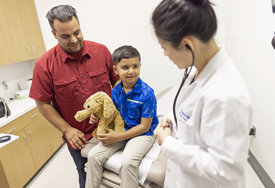 A young boy holds a stuffed dog in a doctor’s office while a doctor and parent look at him.
