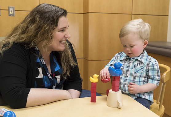 Toddler playing with blocks on a table while female health professional watches and smiles.