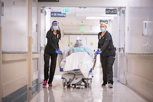 Two providers in black scrubs wheeling a patient down a hallway in a gurney bed.