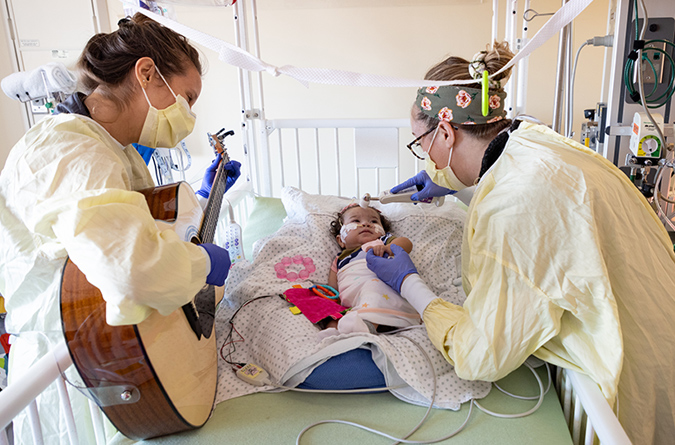A nurse playing guitar while a second nurse tends to a child in the PICU