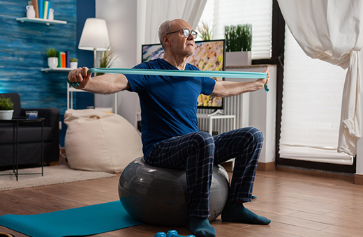 An older man stretches his arms using an exercise band and sits on a yoga ball at home.