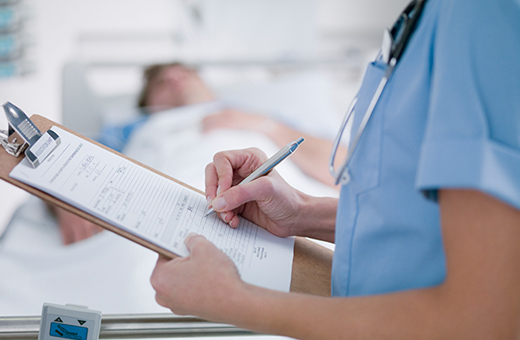 Health care provider making notes on a clipboard in the foreground with patient in hospital bed in the background.