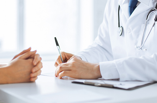 Close up of a desk with a patients hands clasped while a doctor writes on a piece of paper.