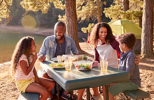 A family sit and talk at a picnic table in front of their tent in the woods. 
