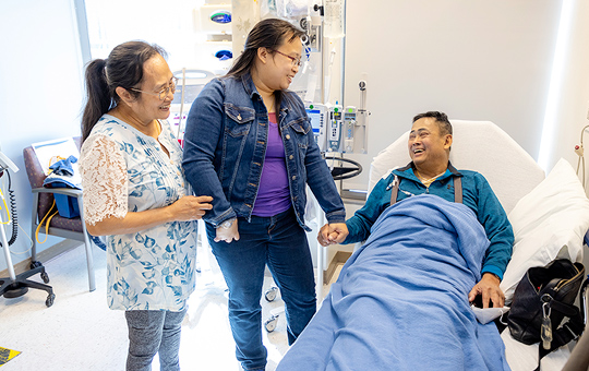 Male patient holding the hand of a family member smiling in a hospital bed.