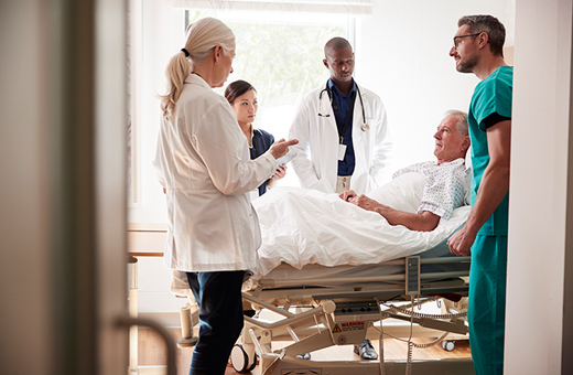 Doctors and nurses stand around an older male patient laying in a hospital bed. 