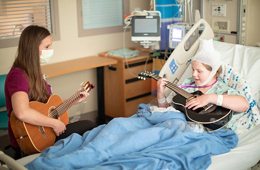 Music therapy specialist playing guitar with a brain surgery pediatric patient in hospital room.