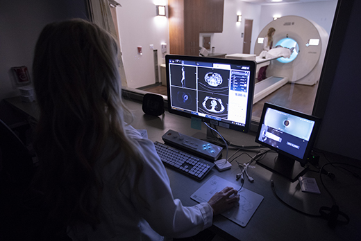 Female radiologist looking at computer screen of images with patient in a PET scan machine in the background