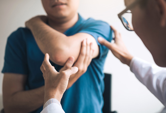 A patient places his hand on his opposite shoulder and stretches his arm as a physical therapist watches.