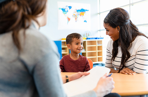 A young child smiles and sits next to his mother while a therapist takes notes.