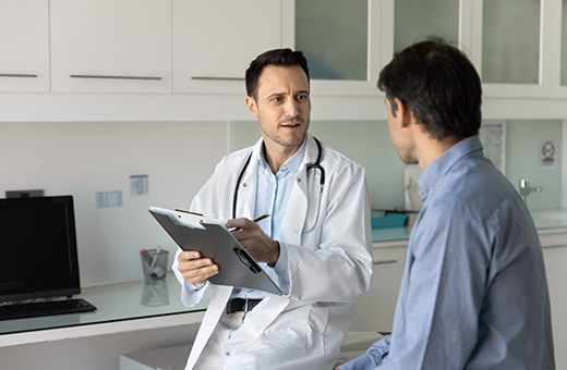 A male doctor holding a clipboard speaks to a male patient.