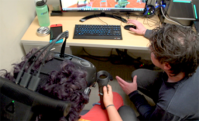 A boy wearing a virtual reality headset is shown by an adult how to use a computer program.