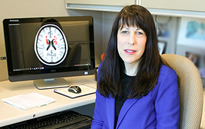 A woman wearing a purple jacket sits next to her computer, which shows a brain scan image.