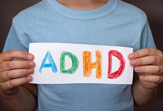 Torso of a young boy in a blue shirt holding a sign that says &#x201c;ADHD&#x201d; in crayon.