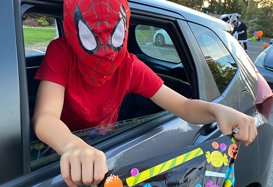 child wearing a spiderman mask reaches out of a window of a gray sedan with a trick-or-treat bag.