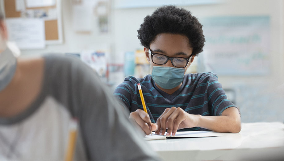 One adolescent boy sits at a school desk, holding a pencil, wearing a surgical mask. 