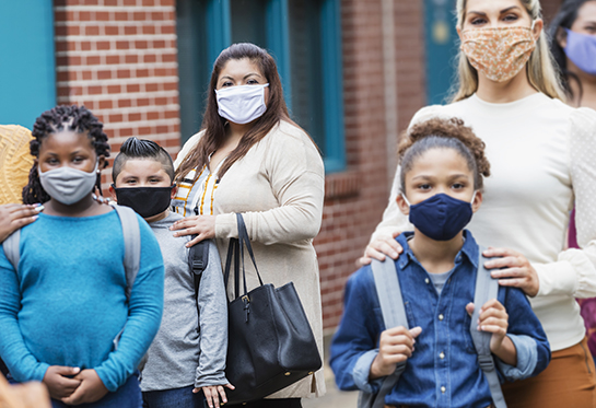 a group of masked diverse parents and their adolescent children stand outside a school