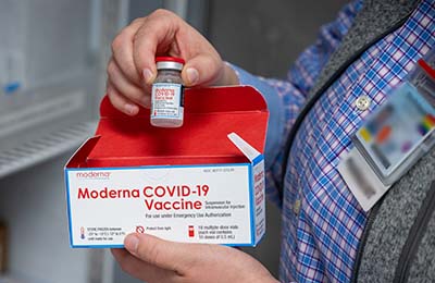 Close-up of a pharmacist’s hand holding a clear bottle of Moderna COVID-19 vaccine with a red label
