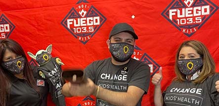 Three people wearing face masks pose in front of a Fuego 103.5 radio station banner.