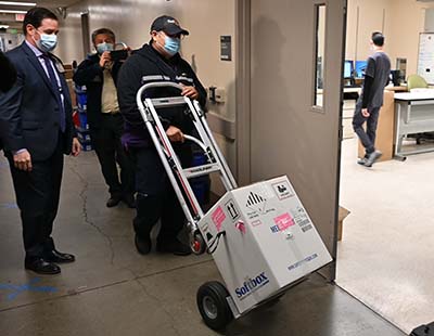 A FedEx delivery driver wheels in a large white box containing Pfizer vaccine, flanked by Chad Hatfield, chief pharmacy officer.
