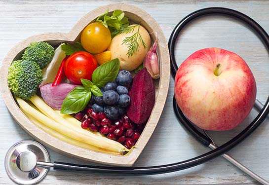 Fruit and vegetables in a wooden heart-shaped box next to a stethoscope and an apple.