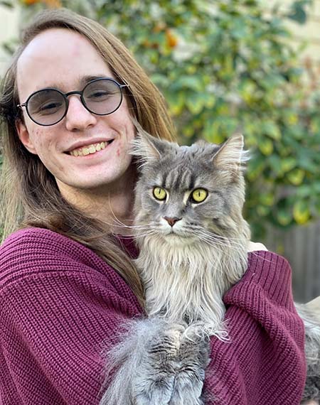 Nick Anderson, wearing a maroon shirt, smiles while holding a fluffy gray cat in his arms, outside. 