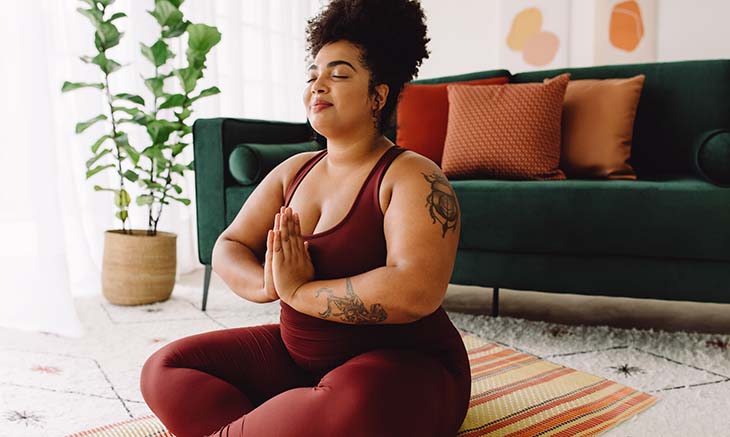 Body positive woman exercising with eyes closed and hands joined while sitting cross legged in living room