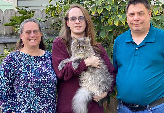 A family of three, mom, dad and teenage son, stand outside in a garden, while the teenage son holds a fluffy gray cat.