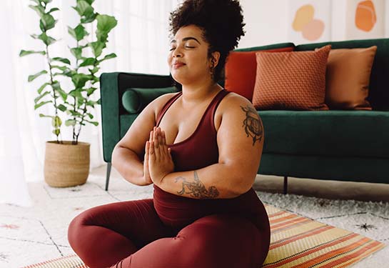 Body positive woman exercising with eyes closed and hands joined while sitting cross legged in living room.